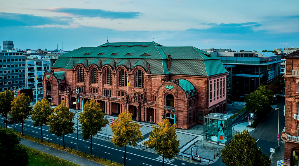 Der Rosengarten Mannheim - Historisches Gebäude mit roter Backsteinfassade und grünem Dach, umgeben von Bäumen. Die großen, bogenförmigen Fenster verleihen dem Bauwerk einen klassischen Charakter. Im Hintergrund kontrastieren moderne Gebäude die traditionsreiche Architektur.