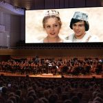 Ein großes Orchester spielt auf einer Bühne vor einem vollen Konzertsaal. Im Hintergrund ist eine Leinwand mit einem Bild zweier Personen