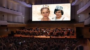 Ein großes Orchester spielt auf einer Bühne vor einem vollen Konzertsaal. Im Hintergrund ist eine Leinwand mit einem Bild zweier Personen