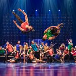 Tanzensemble bei einer farbenfrohen Bühnenperformance mit akrobatischen Sprüngen und blauer Lichtkulisse