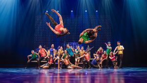 Tanzensemble bei einer farbenfrohen Bühnenperformance mit akrobatischen Sprüngen und blauer Lichtkulisse