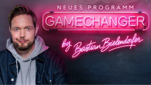 Ein Mann in Jacke und grauem Hoodie steht vor einem Neon-Schild. Das Neon-Schild trägt die Aufschrift 'NEUES PROGRAMM GAMECHANGER by Bastian Bielendorfer'.