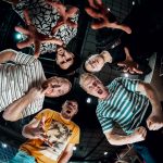 Fünf Personen stehen in einem Kreis und schauen nach unten in die Kamera. Die Perspektive ist von unten aufgenommen, sodass die Gesichter und Oberkörper der Personen zu sehen sind. Alle Personen haben unterschiedliche Gesichtsausdrücke und Gesten, was auf eine fröhliche oder ausgelassene Stimmung hindeutet. Im Hintergrund sind Lichter und Teile eines Gebäudes zu erkennen.