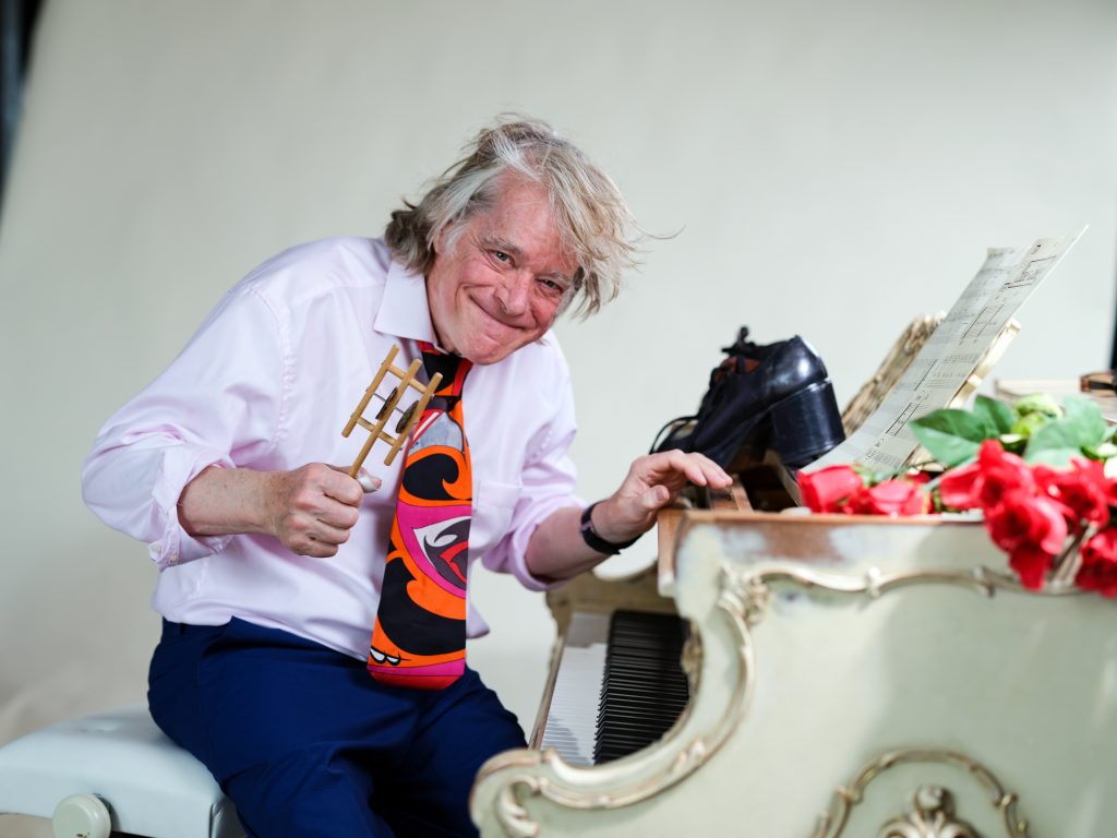 Ein älterer Mann mit grauem Haar sitzt an einem weißen Klavier. Er trägt ein weißes Hemd, eine bunte Krawatte und eine blaue Hose. In seiner rechten Hand hält er einen goldenen Gegenstand, während seine linke Hand auf den Klaviertasten ruht. Auf dem Klavier liegen Notenblätter und rote Blumen.