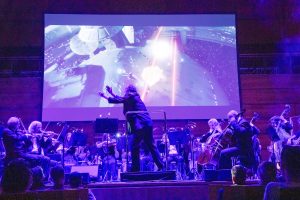 Dirigent dirigiert Orchester auf der Bühne vor dem Leinwand, Publikum sieht zu
