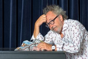 Ein nachdenklicher Mann sitzt an einem Tisch, stützt den Kopf auf die Hand und blickt auf eine zerknüllte Zeitung vor sich. Im Hintergrund hängen dunkelblaue Vorhänge