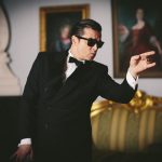 Person in elegantem schwarzen Anzug mit Fliege und schwarzer Sonnenbrille steht in einem stilvoll eingerichteten Raum mit antiken Möbeln und zwei gerahmten Porträts an der Wand. Die Person zeigt mit einer Hand zur Seite