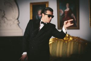Person in elegantem schwarzen Anzug mit Fliege und schwarzer Sonnenbrille steht in einem stilvoll eingerichteten Raum mit antiken Möbeln und zwei gerahmten Porträts an der Wand. Die Person zeigt mit einer Hand zur Seite