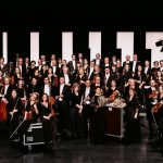 Gruppenfoto eines großen Orchesters auf einer Bühne mit formell gekleideten Musikerinnen und Musikern sowie verschiedenen klassischen Instrumenten