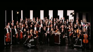 Gruppenfoto eines großen Orchesters auf einer Bühne mit formell gekleideten Musikerinnen und Musikern sowie verschiedenen klassischen Instrumenten